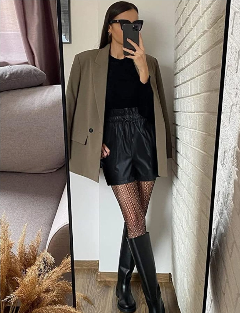 Woman standing with one leg over the other in a army green blazer hanging over her shoulders.  She paired the blazer with a black long sleeve t-shirt and faux leather shorts with sexy opaque tights and black knee length boots.  She made the look fabulous with black sunglasses.